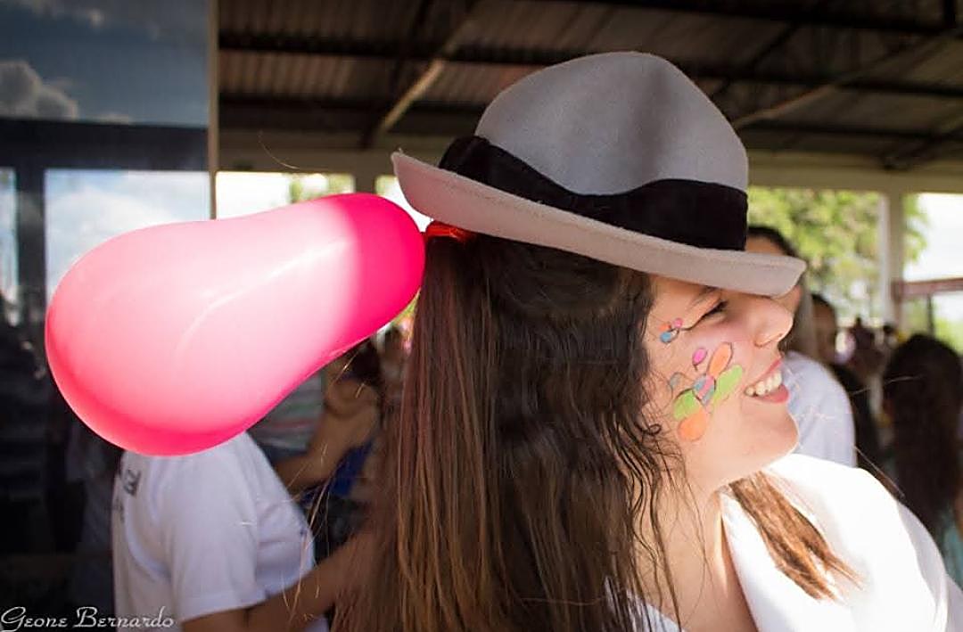 Adriana com um chapéu e uma bexiga rosa presa atrás dele, simulando o penacho de Cyrano de Bergerac, o rosto pintado, sorrindo durante uma contação de histórias ao ar livre