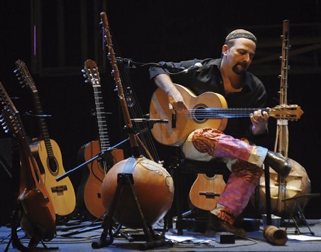 Carlinhos Antunes sentado no palco, tocando violão, cercado por violas, violões, uma kora e instrumentos de percussão apoiados em estantes
