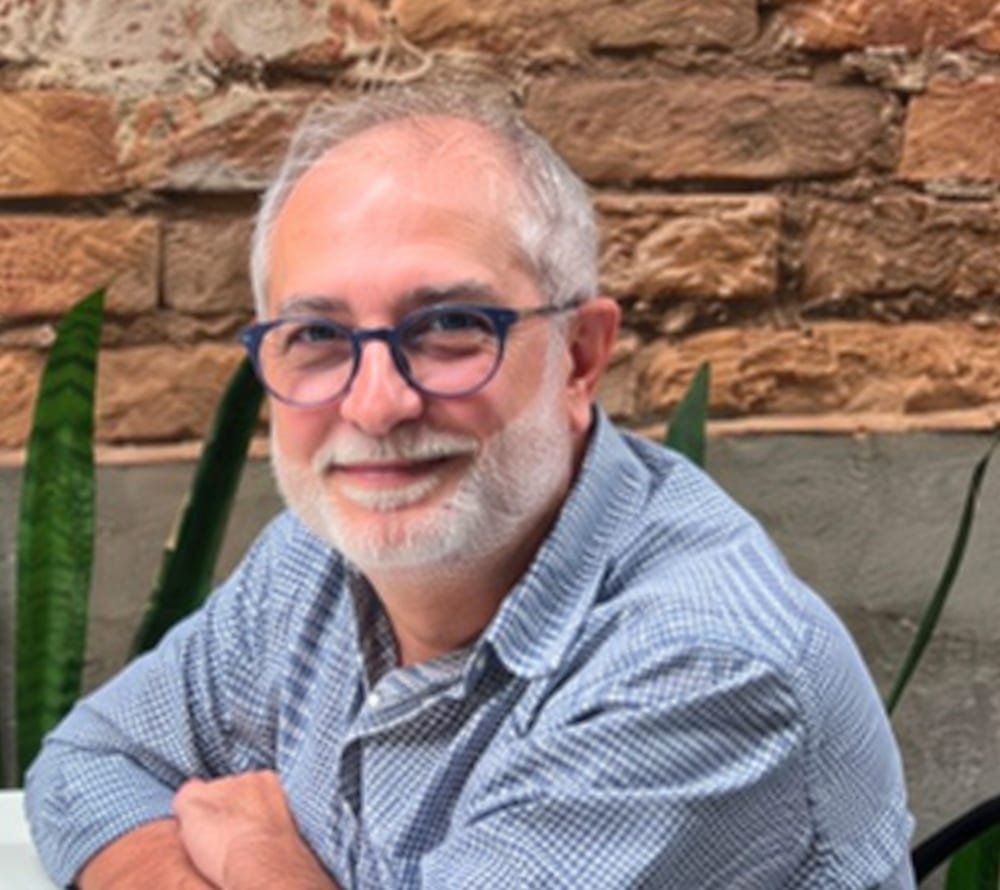Retrato de Claudinei José Pires: homem de cabelos e barba grisalhos, de óculos e camisa xadrez azul-clara, de braços cruzados, sorrindo, diante de uma parede de tijolos aparentes