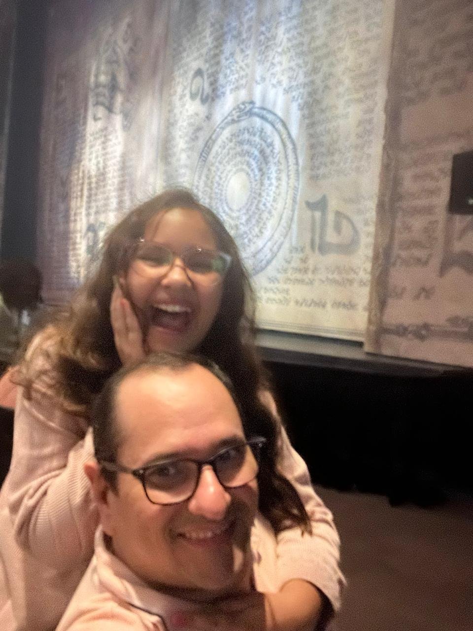 Selfie de Jairo e Elis: ele sorri em primeiro plano e ela, atrás, ri com a mão no queixo; ao fundo, o telão do palco reproduz páginas manuscritas de um grimório