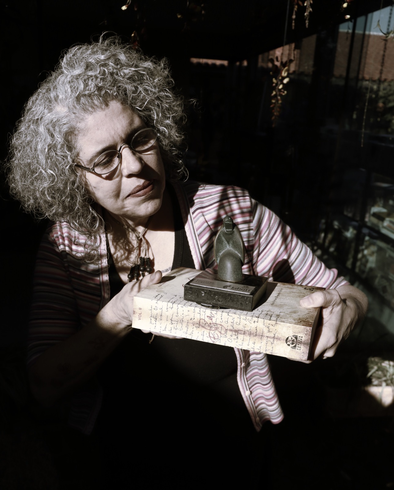 Nima Spigolon segura, sob a luz, o troféu do Prêmio Jabuti — uma pequena escultura de jabuti em bronze — apoiado sobre o volume II do seu livro, cuja capa reproduz um manuscrito