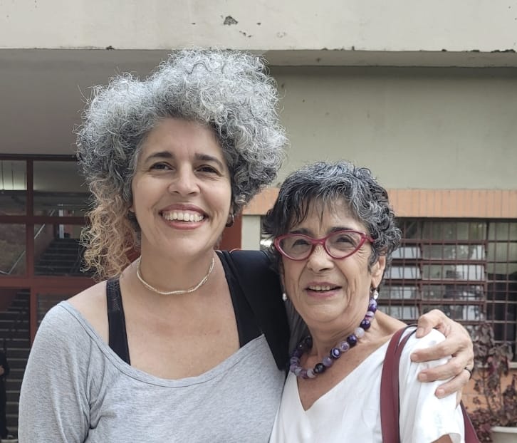 Nima Spigolon, à esquerda, abraça Madalena Freire; as duas sorriem, ao ar livre, em frente a um prédio universitário