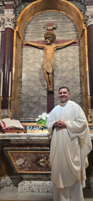 Padre Douglas de vestes brancas, sorrindo, segurando um ramo de flores diante de um altar com a imagem de Cristo crucificado