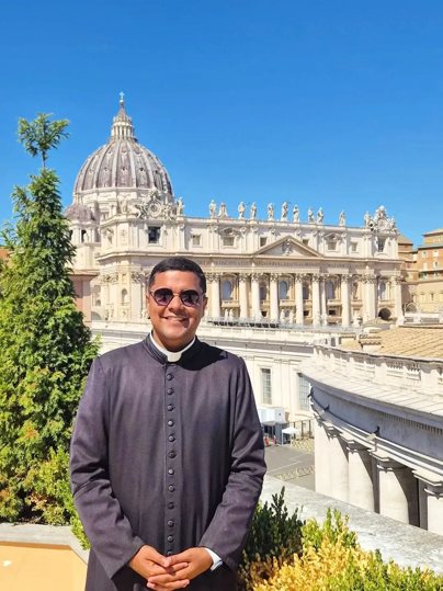 Padre Douglas, jovem, de batina preta e colarinho clerical, usando óculos de sol e sorrindo, diante da Basílica de São Pedro, no Vaticano, sob céu azul