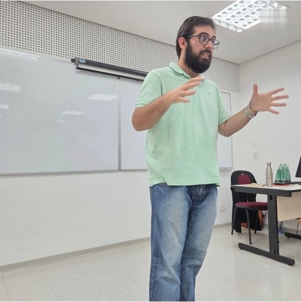 Rodney Amador em pé, de camisa polo verde-clara, gesticulando com as duas mãos diante de uma lousa branca em sala de aula