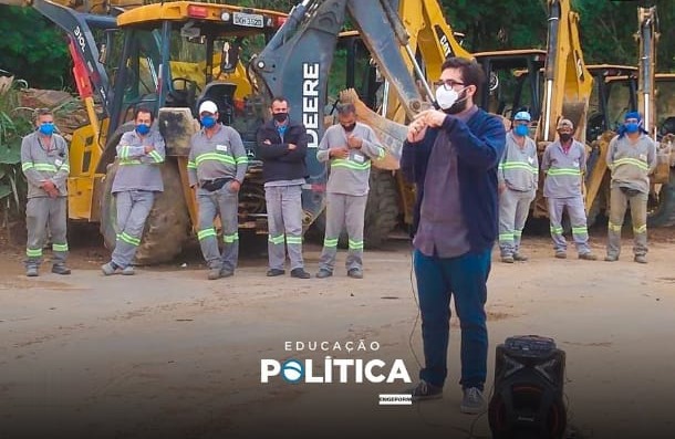 Rodney Amador, de máscara, fala ao microfone diante de operários uniformizados, com faixas refletivas, alinhados em frente a retroescavadeiras amarelas