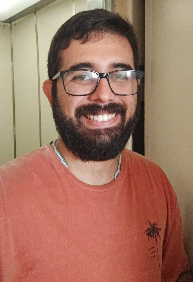 Selfie de Rodney Amador sorrindo, de óculos, barba, camiseta laranja e colar de miçangas