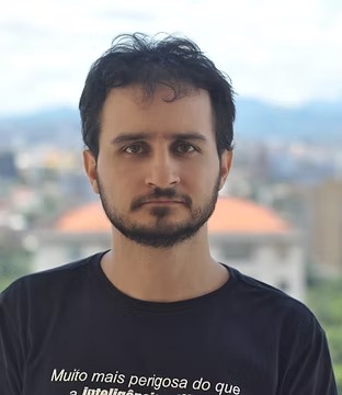 Retrato de Rômulo Soares Valentini, de cabelo escuro e barba, com camiseta preta estampada, ao ar livre, com a cidade e as montanhas desfocadas ao fundo